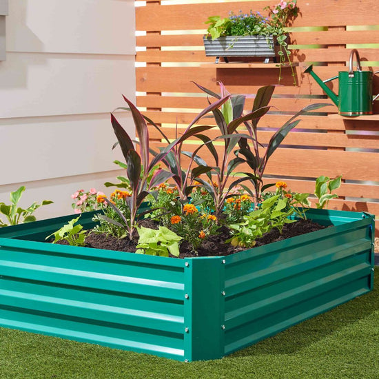 A teal green metal raised garden bed filled with green and burgundy plants, set on artificial grass in front of a wooden slat wall with a green watering can and hanging planter visible.