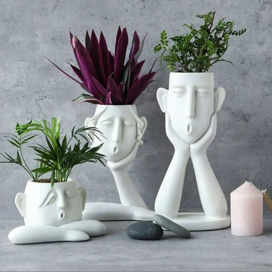Three white ceramic planters shaped like stylized human faces arranged on a gray surface against a textured gray wall. The planters hold green and burgundy plants, with a small pink candle and dark stones beside them.