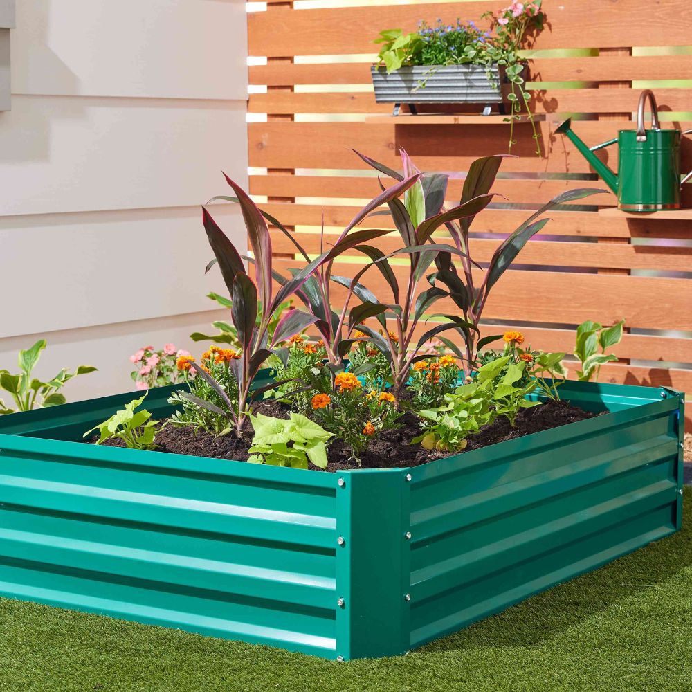 A teal green metal raised garden bed filled with green and burgundy plants, set on artificial grass in front of a wooden slat wall with a green watering can and hanging planter visible.